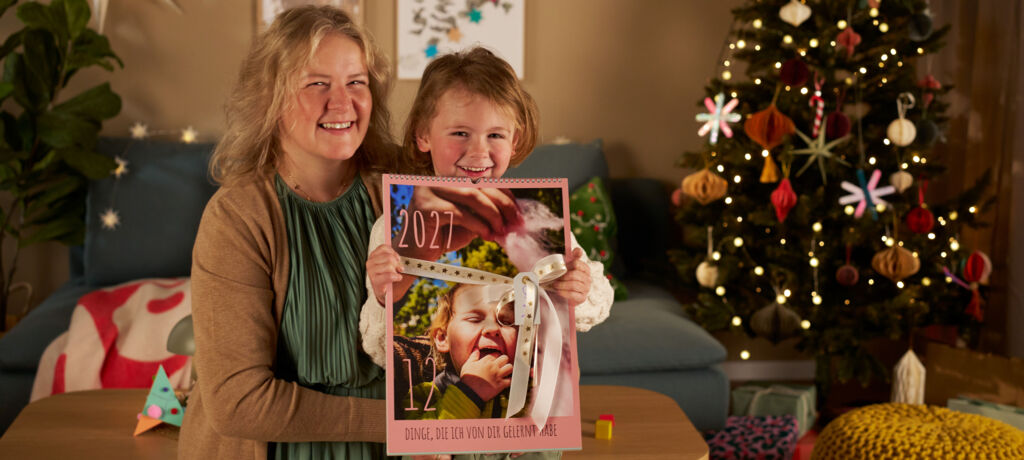 Ein Kind uns seine Mama stehen in einem Wohnzimmer neben einem geschmückten Weihnachtsbaum. Der Junge hält einen Fotokalender in die Kamera.