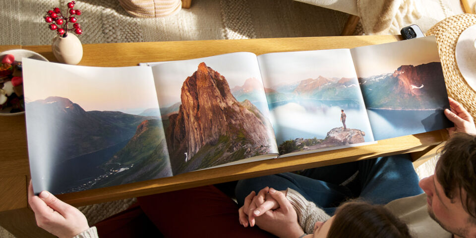 Zwei Personen betrachten ein geöffnetes CEWE FOTOBUCH auf einem Holztisch, das eine Berglandschaft zeigt.