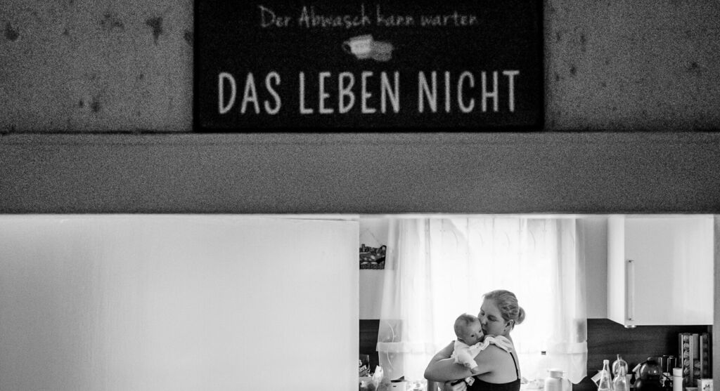 Ein kleines Schild mit der Aufschrift „Der Abwasch kann warten, das Leben nicht“. Im Hintergrund ist eine Mutter mit Neugeborenem.