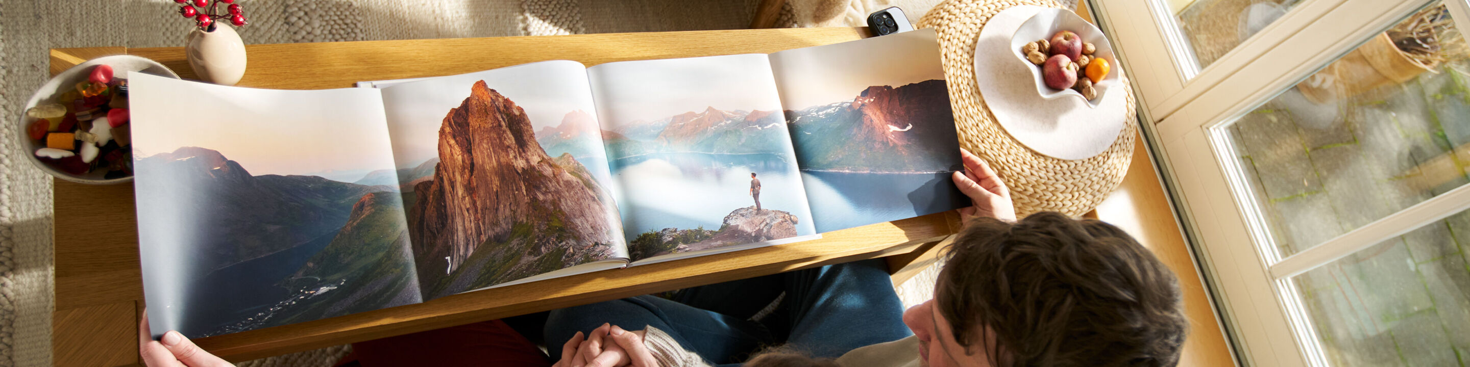 Zwei Personen betrachten ein geöffnetes CEWE FOTOBUCH auf einem Holztisch, das eine Berglandschaft zeigt.