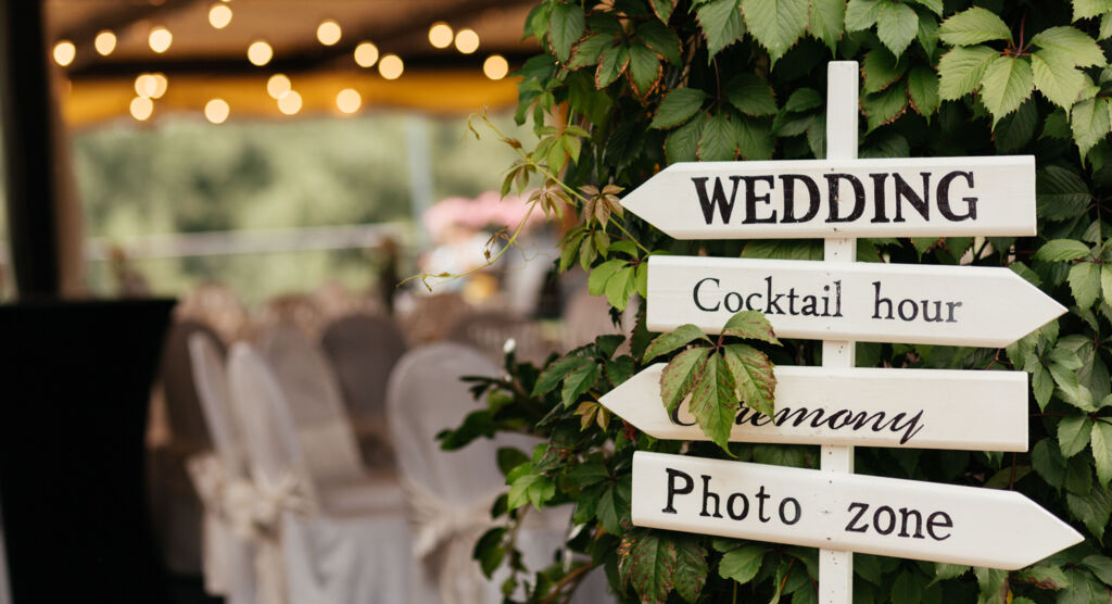 Wegweiser bei einer Hochzeitsfeier: Pfeile zeigen zum Cocktailbereich, zur Zeremonie und zur Fotoecke. Im Hintergrund sind geschmückte Stühl