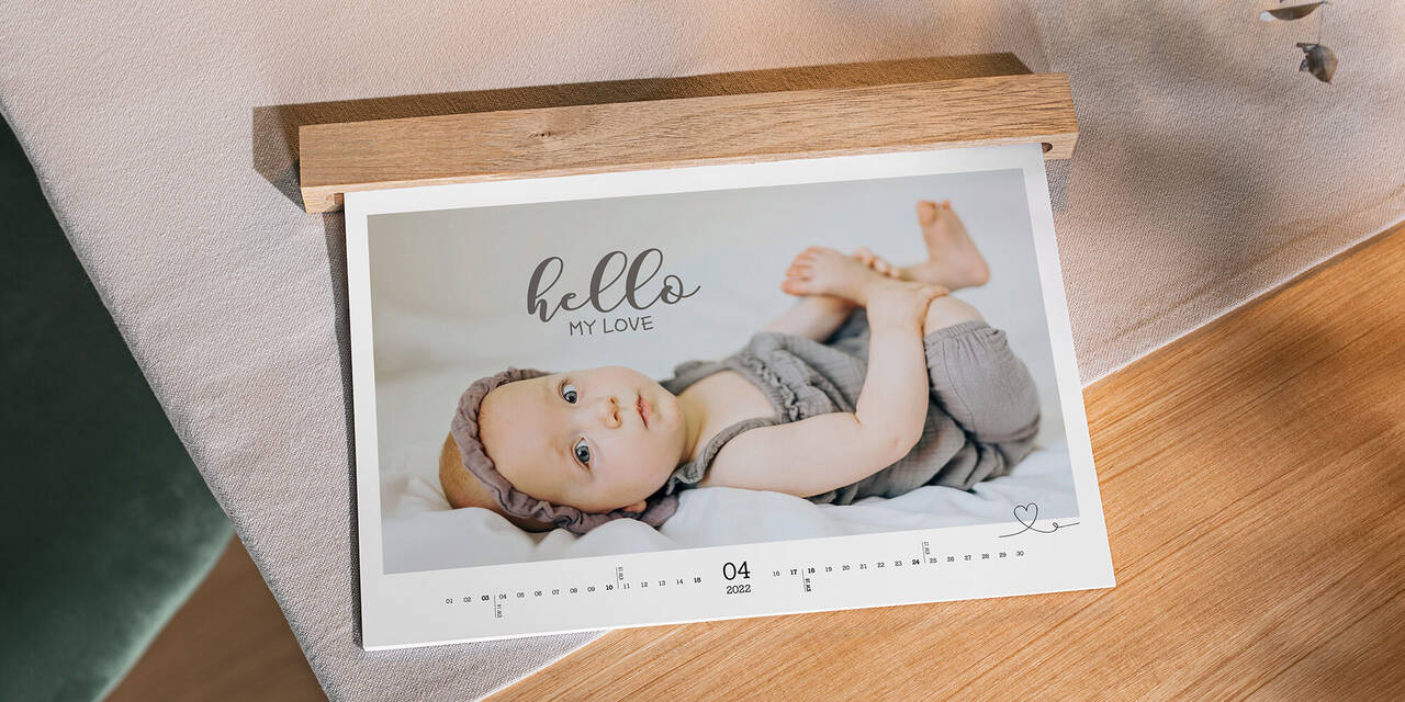 Auf einem Tisch aus Holz liegt ein Wandkalender mit Babymotiven im Querformat. Er zeigt den Monat April und hat eine Holzleiste, die die Ringbindung verdeckt.
